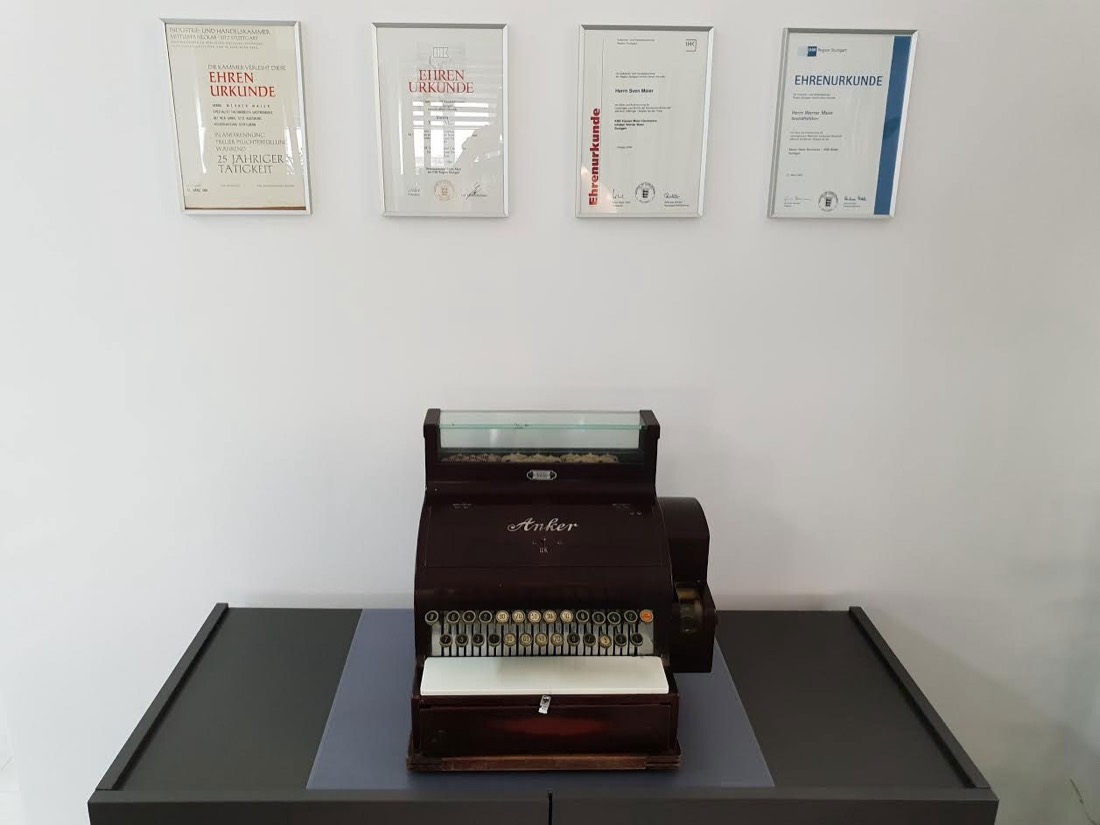 Historische Registrierkasse im Ausstellungsraum von Kassen Maier Electronics, darüber vier gerahmte Urkunden an der Wand